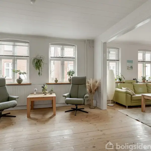 To lænestole er placeret foran store vinduer med planter på karmen i en lys stue. Ved siden står en grøn sofa med et sofabord. Trægulve og hvide vægge skaber en hyggelig atmosfære.