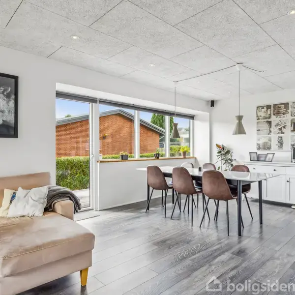 En moderne stue med en beige sofa til venstre, et spisebord med fem stole i midten, og store vinduer, der afslører en udsigt til en murstensbygning udenfor.