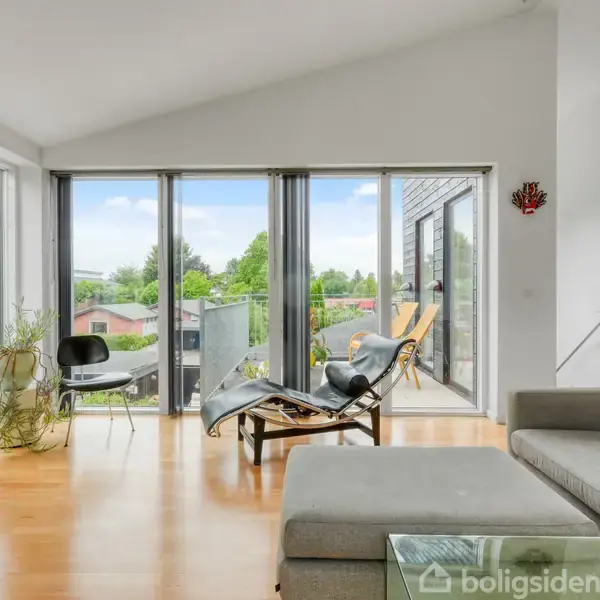 En lys stue med trægulv, hvor en grå sofa står ved en glasbord. Store vinduer og altandøre slipper sollys ind. En stol og planter står nær en udgang til balkonen.