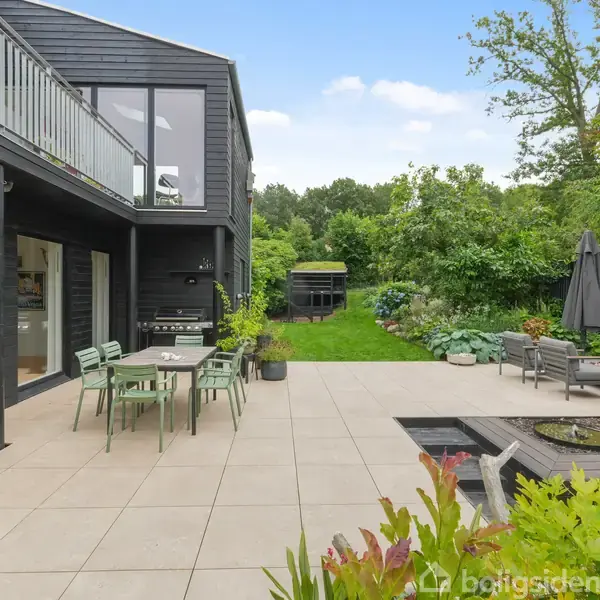 Sort træhus med store vinduer. Udendørs terrasse med bord og stole. Haven har græsplæne og planter, omgivet af træer. Borde og stole på terrassen, solrig dag.