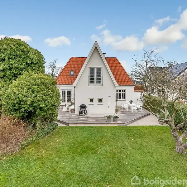 Et hvidt hus med rødt tag står i en grøn have. Foran huset er en terrasse med havemøbler. Store buske og et træ omkranser haven.