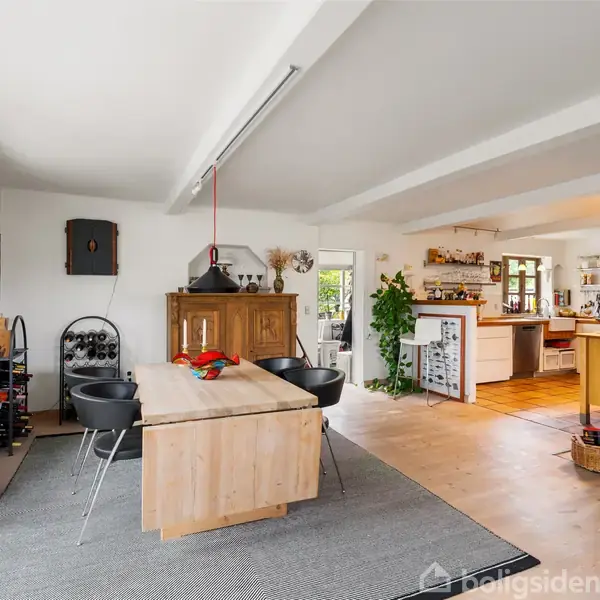 Træbord med sorte stole står i spisestue. Pejs er tændt ved siden af, mens køkkenet med hylder og planter ses i baggrunden. Stemningen er hjemlig og lys.