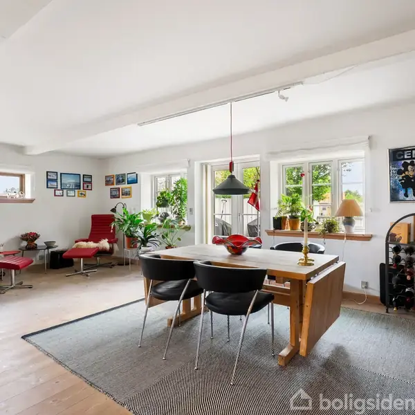 Spisebord med tre stole omkring, i en lys stue. Rummet indeholder også en rød lænestol, planter, kunst på væggene, og store vinduer med udsigt til grønne omgivelser.