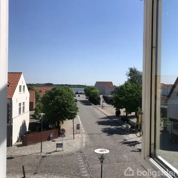 Udsigt fra et vindue. Gade med træer og huse med røde tage, førende mod en flod eller et vandløb i det fjerne. Solrig, blå himmel.