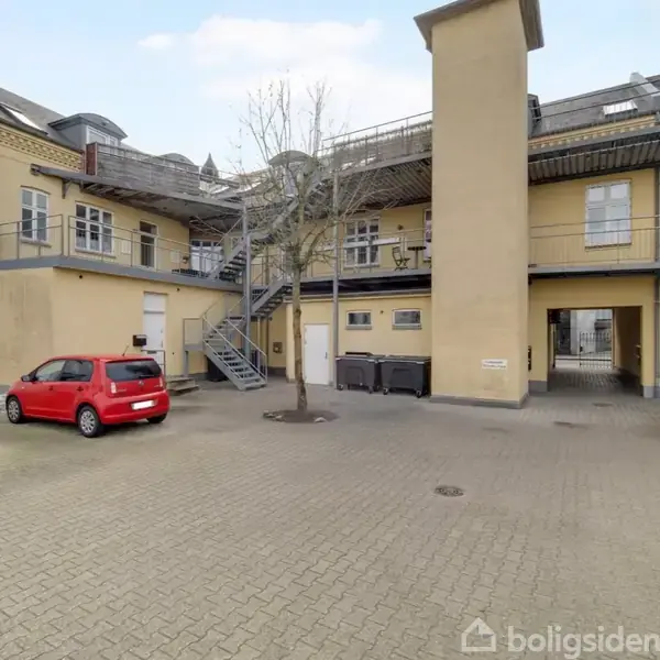 En gårdsplads med parkerede biler, hvor en rød bil holder til venstre. En murstensbygning med trapper ses i baggrunden, og et træ står i midten.