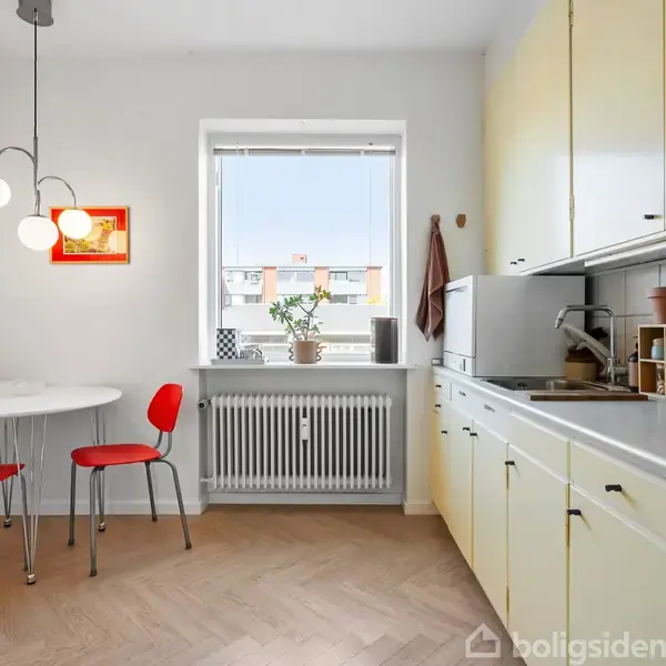Et lyst køkken med hvide skabe, et spisebord med røde stole ved et vindue. Plante og kogebog på hylden. Lampen hænger fra loftet. Ingen tekst fundet.