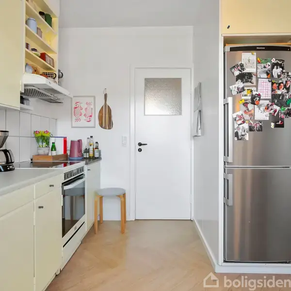Køkken med lyse skabe og en stål køleskab fyldt med magneter og fotos. Der er en ovn, kaffemaskine og diverse køkkenredskaber på bordpladen. En dør fører til et andet rum.
