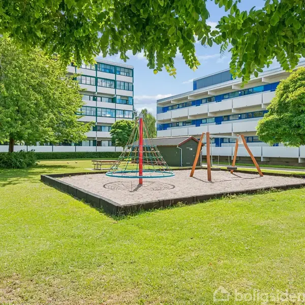 Legeplads med klatrestativ og gynger placeret i sandkasse midt i grøn park omgivet af græsplæne og træer. Baggrund med moderne, fleretages lejlighedsbygninger under en blå himmel.
