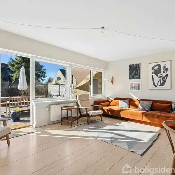 En lys stue har en brun sofa, ved siden af et stort vindue, der åbner ud til en terrasse med havemøbler. Væggene er dekoreret med minimalistiske kunstværker.