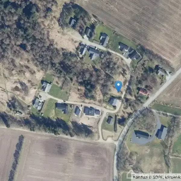Et satellitbillede viser en række boliger beliggende i et skovklædt område omgivet af marker. En blå markeringspin angiver en specifik placering blandt bygningerne.