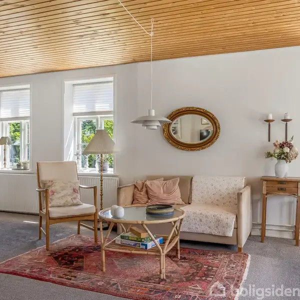 En stue med en beige sofa og stol, et rundt sofabord med bøger og en lampe over. Trævægge og -loft, rødt tæppe på gulvet, lys fra vinduer giver rummet en lys atmosfære.