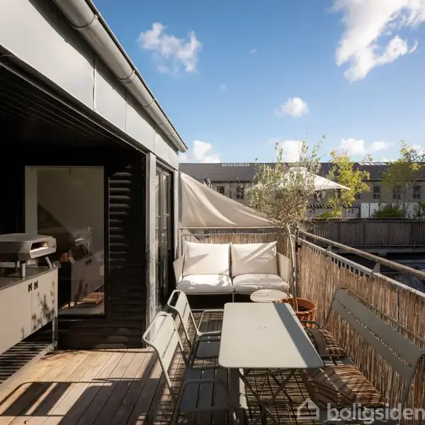 Udendørs terrasse med træbord og stole, loungemøbler og planter. Terrasse adskilt med hegn, udsigt til nabobygninger. Sollys skaber en lys og indbydende atmosfære under en blå himmel med få skyer.