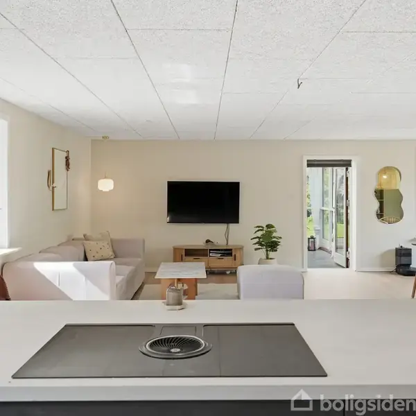 Stue med sofa og sofabord foran fladskærms-tv på væggen. Stor ø i forgrunden indeholder kogeplade. Spisebord med stole ved siden af vinduer. Indgang til terrasse i baggrunden.
