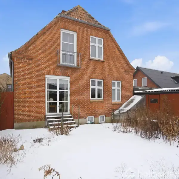 En murstenshus med store vinduer står stille i et sneklædt haveområde, omgivet af nabobygninger og klare, blå himmel.