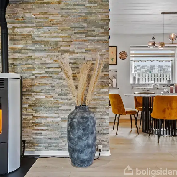 Brændeovn opvarmer rummet mod stenmur, mens en stor vase med tørrede græsser står ved siden af. Bagved ses et spiseområde med orange stole og moderne belysning.