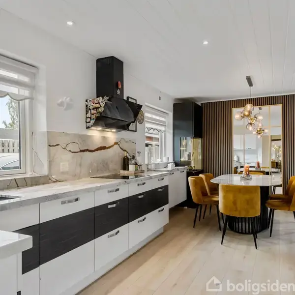 Køkken med moderne design har hvide og sorte skabe, marmor-bordplade og kogeplade ved vindue. Rundt spisebord med gule stole står i baggrunden nær dekorativ lysinstallation. Træpanelvæg i baggrunden.
