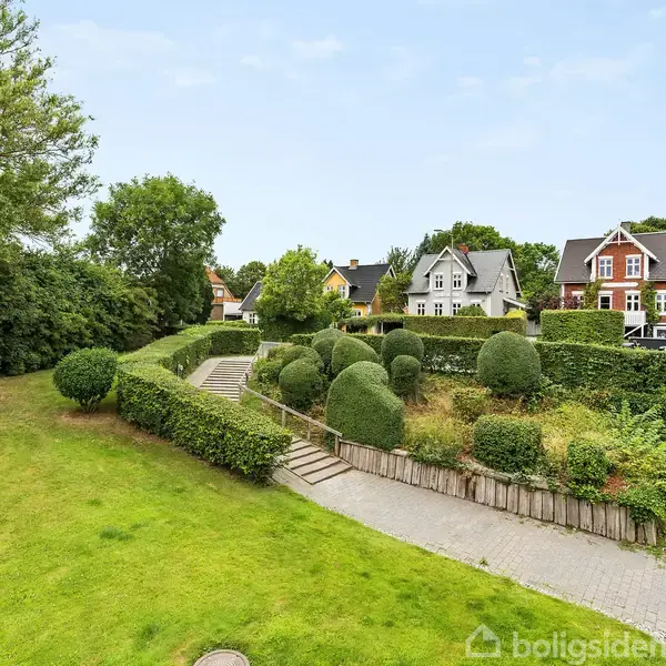 Grønne buske og træer omkranser en velplejet have med en flisebelagt sti, der fører til flere maleriske huse i baggrunden. En blå himmel hænger over landskabet.