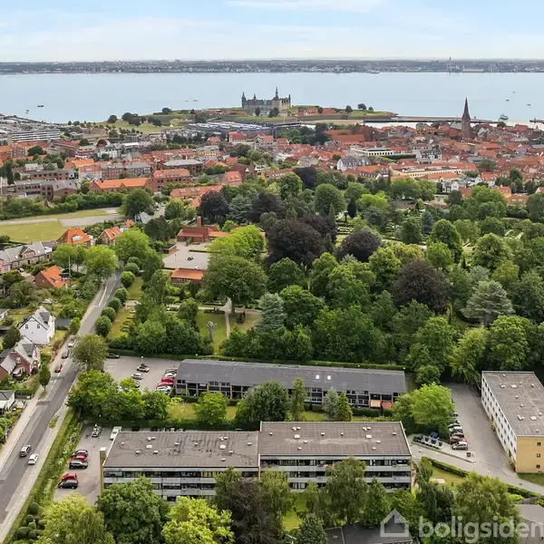 Luftfoto af boligblokke omgivet af træer; i baggrunden ses en by med røde tage og en kirke, samt vand og en stor bygning ved kysten.