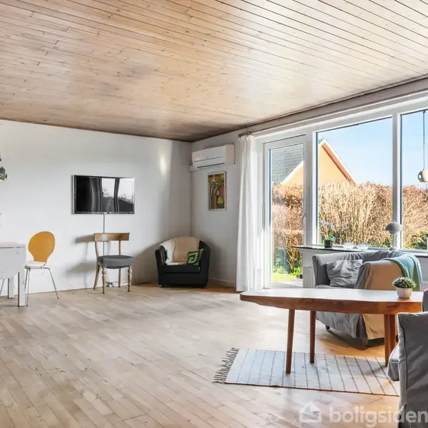 Stort, lyst stueområde med trægulv og træloft. To lænestole og et rundt bord mod venstre væg. Vinduer til højre viser en have. En sofa står rundt om et træbord. Ingen tekst synlig.