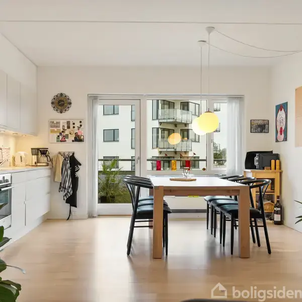 Et moderne køkken med en spisebord med sort stole, omgivet af hvide vægge dekoreret med kunstværker. En stor glasdør fører til en balkon med udsigt over andre bygninger.