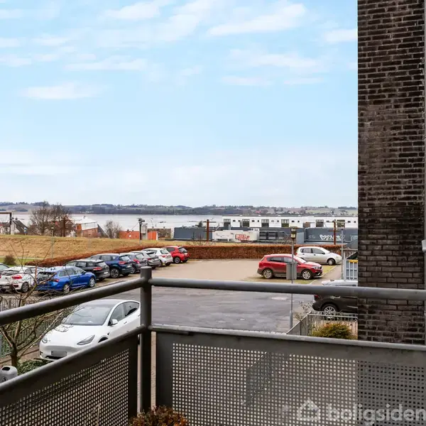 En altan med metalgelænder vender ud mod en parkeringsplads med biler. I baggrunden ses en udsigt over vandet og en by i det fjerne under en blå himmel.