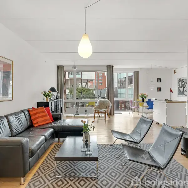 Stue med sort lædersofa og sofabord med blomster, placeret på mønstret tæppe. Tv er monteret på væggen. Store vinduer og spisebord i baggrunden skaber rummelighed.
