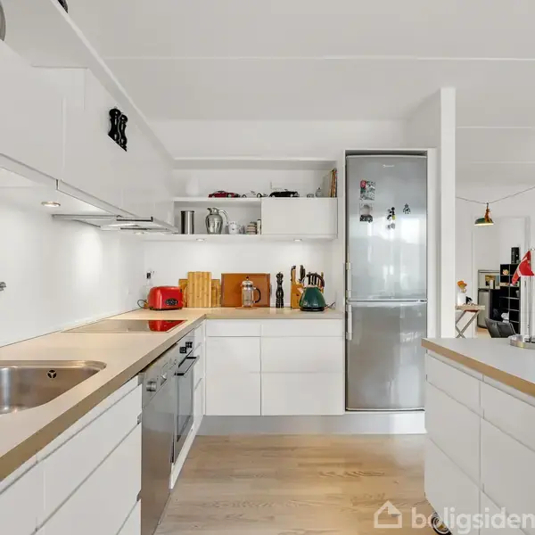 Hvidt køkken med moderne, enkeltsidet indretning. Vask og komfur er placeret langs bordpladen med indbyggede skabe. Et stort køleskab står for enden. Naturligt lys fra opholdsstue.
