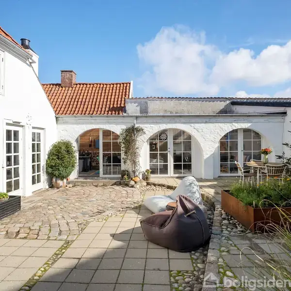 Hvidmalet gårdhave med brosten og fliser, byder på siddemøbler og planter. Omkring ligger huse med rødt tegltag, buevinduer og blå himmel i baggrunden.