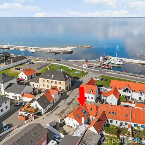 Hus markeret med rød pil ligger i et kvarter med røde taghuse ved en havn. Både ligger fortøjet, og roligt vand ses i baggrunden.