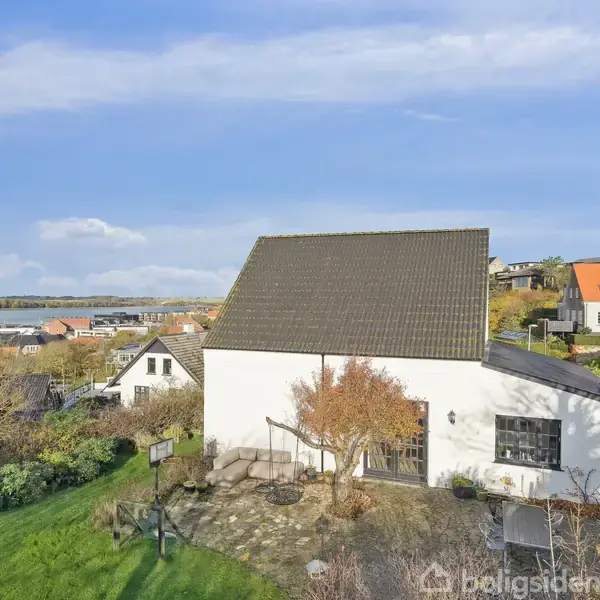 Hvidt hus med mørkt tag står i en grøn have med træer. Omkring ligger andre huse med røde tage. I baggrunden ses en sø eller fjord under en blå himmel.