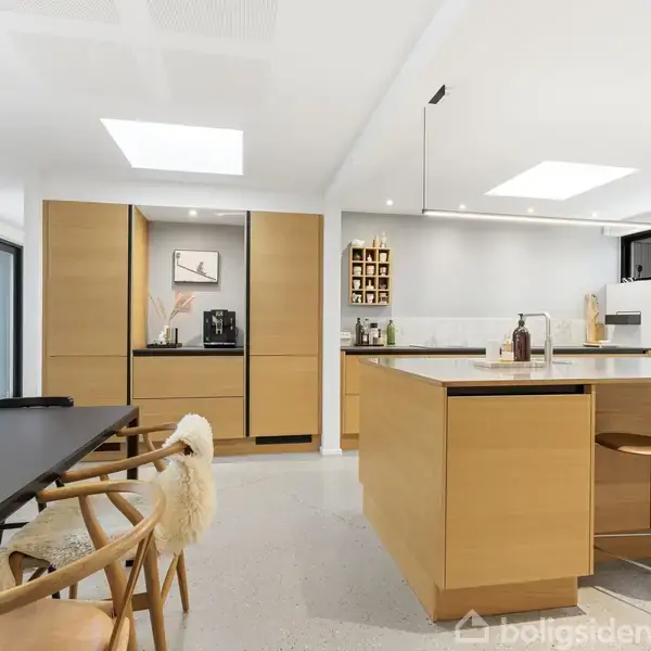 Køkken med træskabe står i moderne indretning, rene linjer og lyse farver. Køkkenø og spisebord i nærheden, omgivet af stole, under naturligt lys fra ovenlysvinduer.