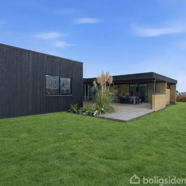 Moderne hus med sorte træpaneler og fladt tag står på en velplejet græsplæne. Foran huset er der en terrasse med havemøbler omgivet af buske, i en rolig landlig indstilling.