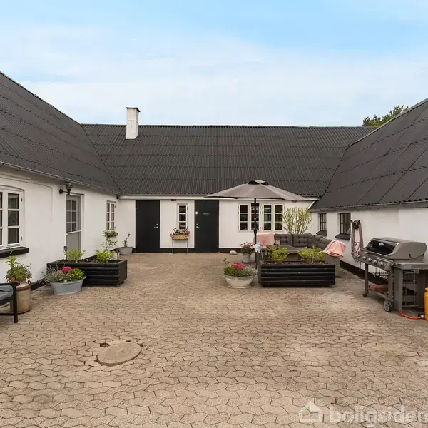 Et gårdmiljø med tre hvidkalkede bygninger. En gårdsplads med flisebelægning har plantekasser, en parasol, bænke og en grill. En dør og to vinduer er synlige midtfor.