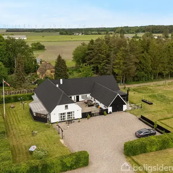 Et hvidt hus med sort tag står ved en grusbelagt indkørsel, omgivet af grønne plæner og træer. En landevej løber langs ejendommen, der ligger i et landligt område.