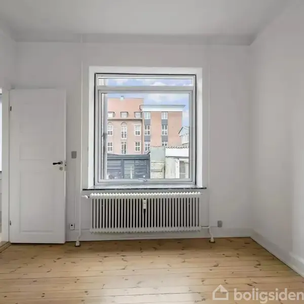 Et tomt rum med lyse trægulve og hvide vægge. Et stort vindue med radiator under giver udsigt til bybygninger udenfor. En åben dør fører til et tilstødende rum.