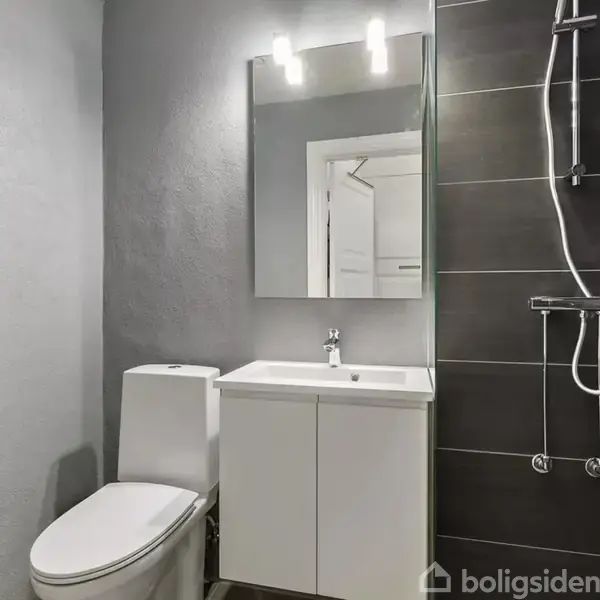 Toilet og håndvask står i et badeværelse med grå vægge. Der er en brusekabine med sorte fliser og en spejl med indbygget lys over vasken.