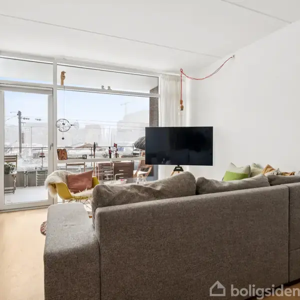 Grå sofa står i en lys stue med trægulv; et fjernsyn hænger på væggen. Store vinduer og glasdør fører ud til en altan med udsigt over byen.