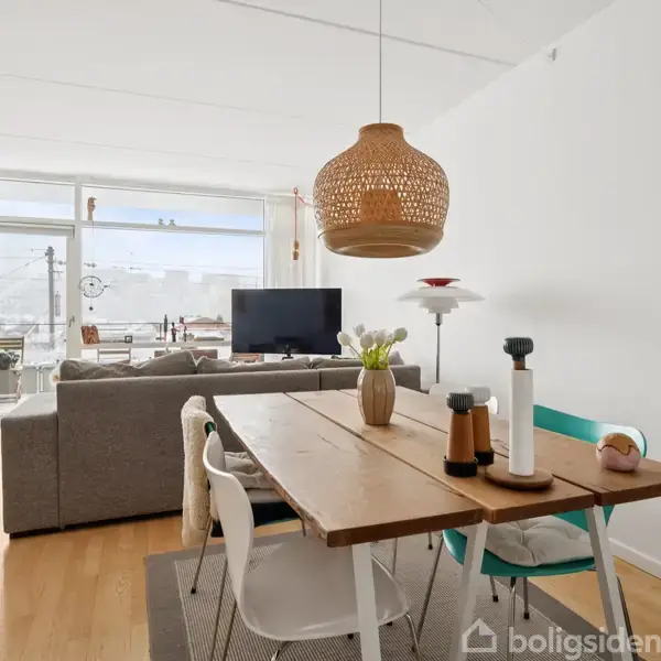 En grå sofa står i stuen, rettet mod et ophængt fjernsyn. Stuen har store vinduer, der fører ud til en balkon med udsigt over byen. Malerier læner sig mod væggen.