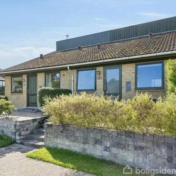 Hus i gule mursten med sorte tagsten er central i billedet. Trætrappe leder op til hoveddøren. Omgivet af en lav mur og frodige buske. Adresse: 101.