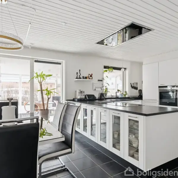 Et moderne køkken med hvide skabe og sorte overflader. Sorte læderstole står ved et spisebord, mens planter pynter rummet. Store vinduer giver udsigt til en terrasse.