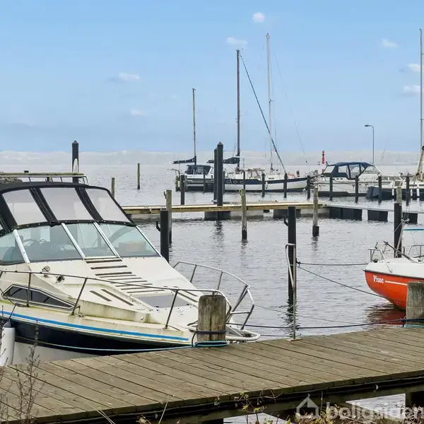 En lystbåd ligger fortøjet ved en bådebro i en marina; flere både er fortøjet i nærheden. Skumsprøjt ses i baggrunden, himlen er let skyet.