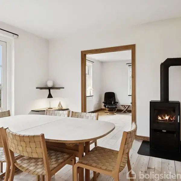 Et rundt spisebord med flettede stole står i et lyst rum med en brændeovn. Glaserede terrassedøre fører ud til en have. I baggrunden ses en stue med stol.