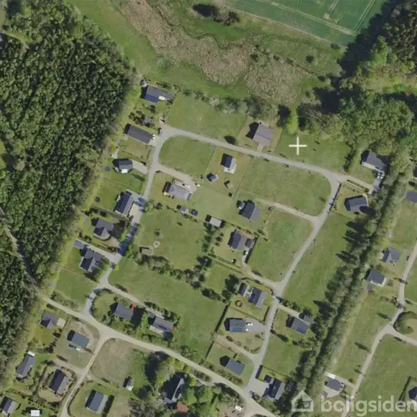 Luftfoto af en landsby viser spredte huse, nogle med haver, placeret langs snoede veje. Omkringliggende område domineret af skove og grønne marker. Plus-tegn ses centralt i billedet.