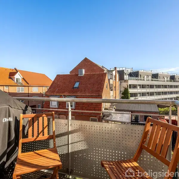 To træstole står på en altan ved siden af en overdækket grill, med udsigt til byhuse med røde tage og en moderne etagebygning i baggrunden under en klar blå himmel.