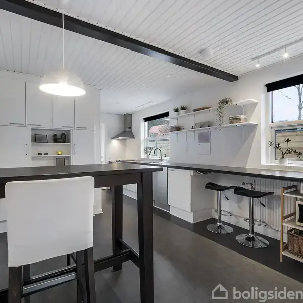 Moderne køkken med hvidt interiør, sort bordplade, to barstole ved en høj bar, og et lille spisebord med en stol. Hylder langs væggen. Store vinduer giver naturligt lys.