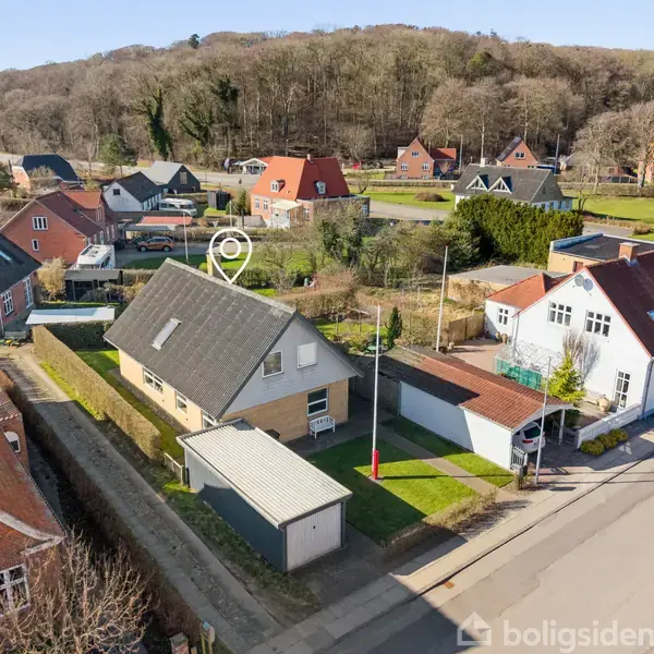 Hus med gråt tag og garage står ved vejen i et boligkvarter. Omgivet af andre huse med røde og grå tage. Baggrund viser skovklædte bakker. Ingen synlig tekst i billedet.