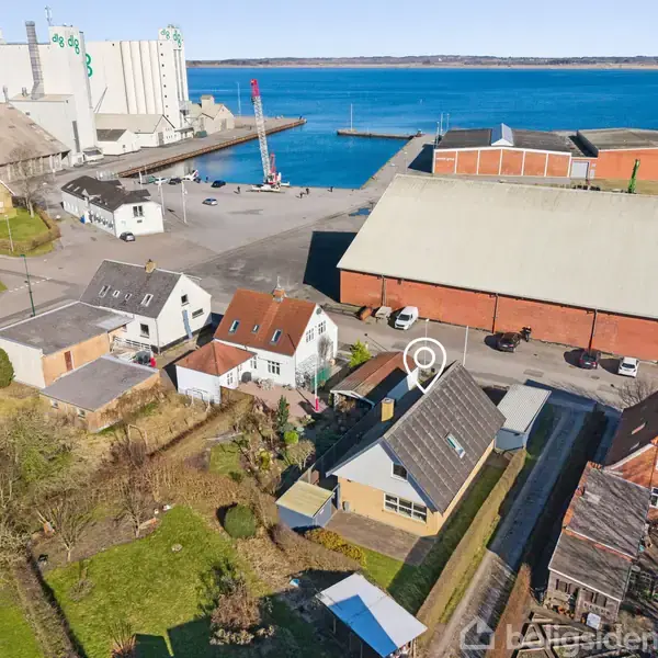 Et hus med mørkt tag og have, placeret tæt på andre bygninger. Baggrunden viser en havn med industrihaller, nogle både og en blå vandflade.