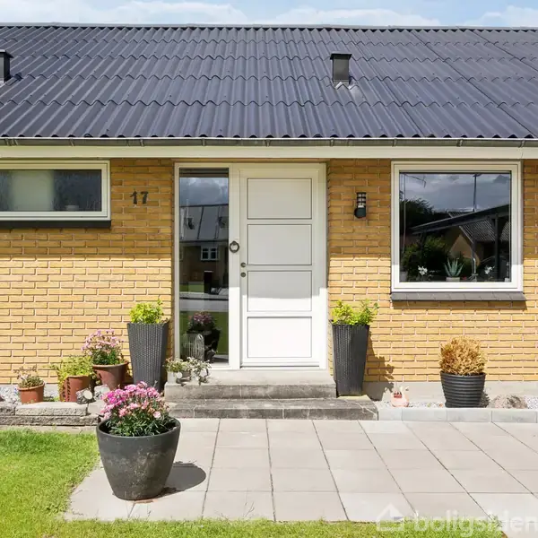 Et gult murstenshus med sort tag har en hoveddør omkranset af blomsterkrukker på flisebelagt terrasse. Husnummer 17 er synligt ved siden af døren.