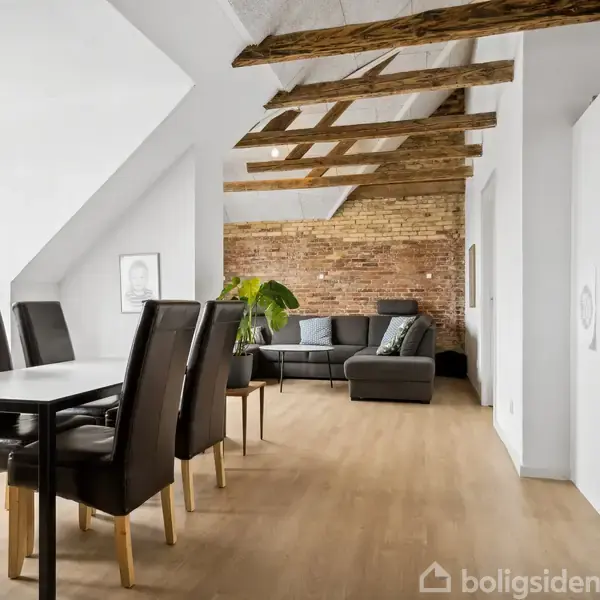 Spisebord med seks stole står i en lys stue med skrålofter. Stuen har en sofa, træbjælker i loftet og en murstensvæg, med et stort vindue til venstre.