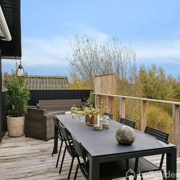 En lang spisebord med stole står på en træterrasse. Bordet er pyntet med blomster og dekorative genstande. Terrassen har udsigt til frodig vegetation og en klar blå himmel.